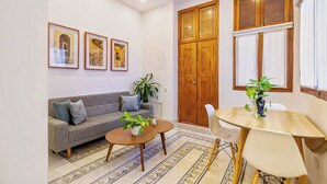 Una sala de estar con sofá, mesa de centro y una mesa de comedor con sillas. Hay cuadros enmarcados en la pared, una puerta de madera grande y una ventana con cortinas.