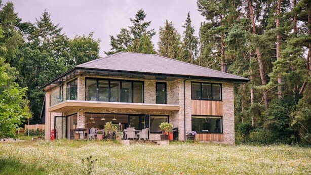 A modern two-story house with a large patio area, surrounded by a field of wildflowers and tall trees. - Stunning new Build by the River (Costessey)