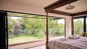 A bedroom with a large window, a four-poster bed, and a view of a garden. - Stunning new Build by the River (Costessey)