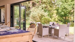 A dining area with a table set for four, surrounded by wicker chairs, and a glass door leading to a garden with trees and shrubs. - Stunning new Build by the River (Costessey)