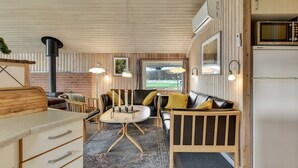 A cozy cabin interior with a wooden ceiling, a dining area with a round table, a sofa with cushions, a kitchen area with a refrigerator, and a large window offering a view of the outdoors. - "Hjalmar" - 400m from the sea by Interhome (Millinge)