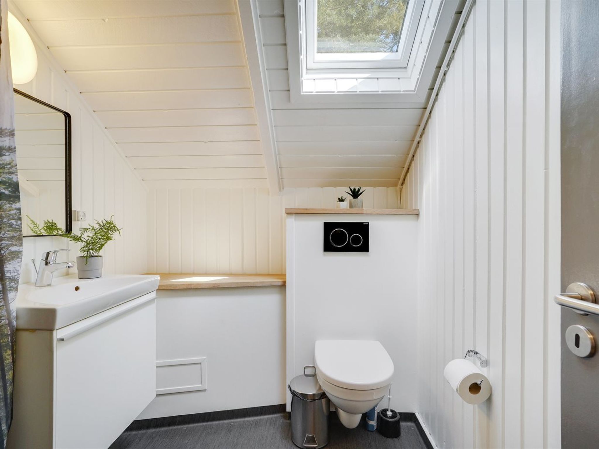 A modern bathroom with a skylight, white walls, a sink, toilet, and a trash bin.
