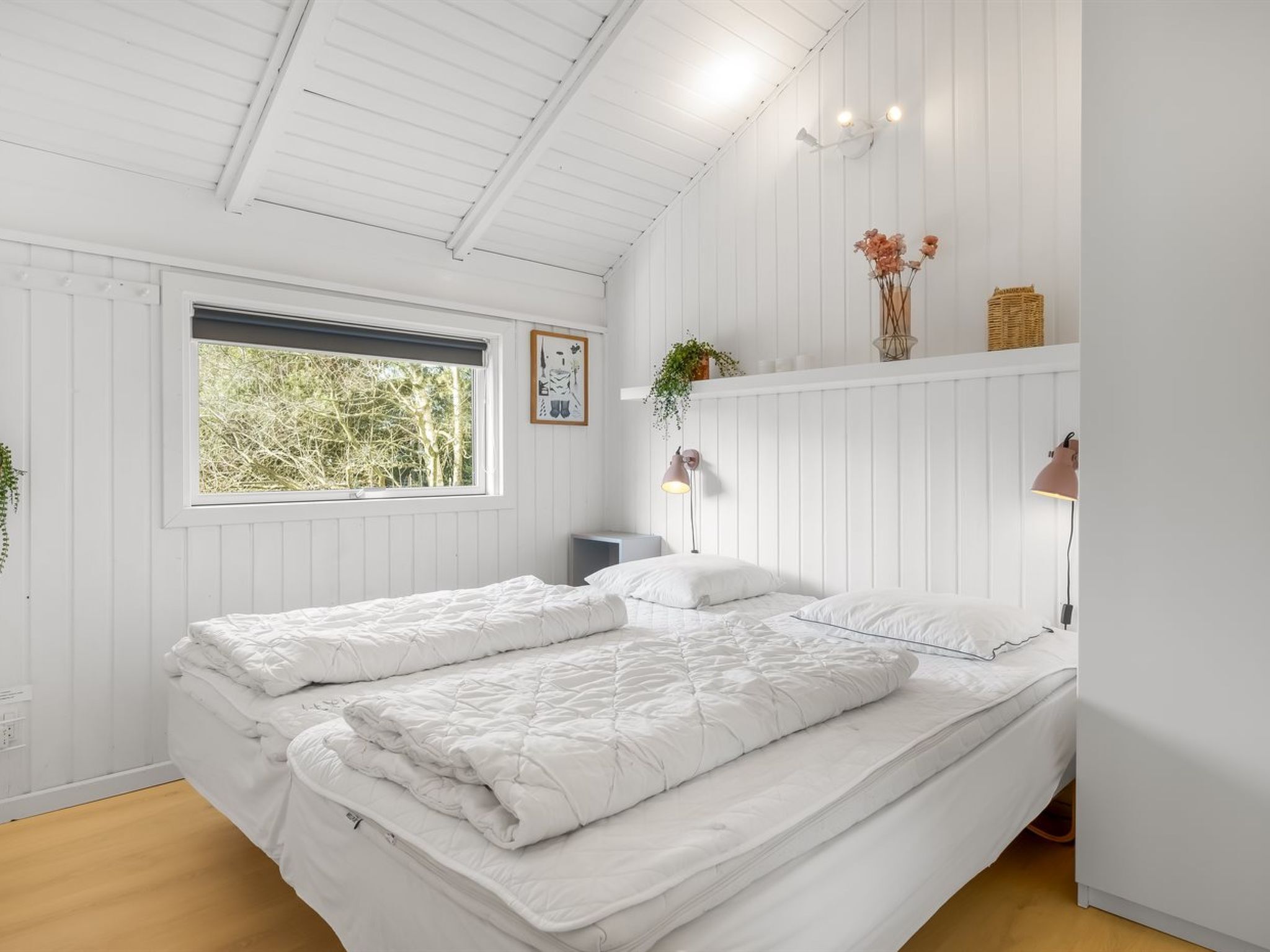A bedroom with a bed, a window, a shelf with plants, and a wall-mounted lamp.