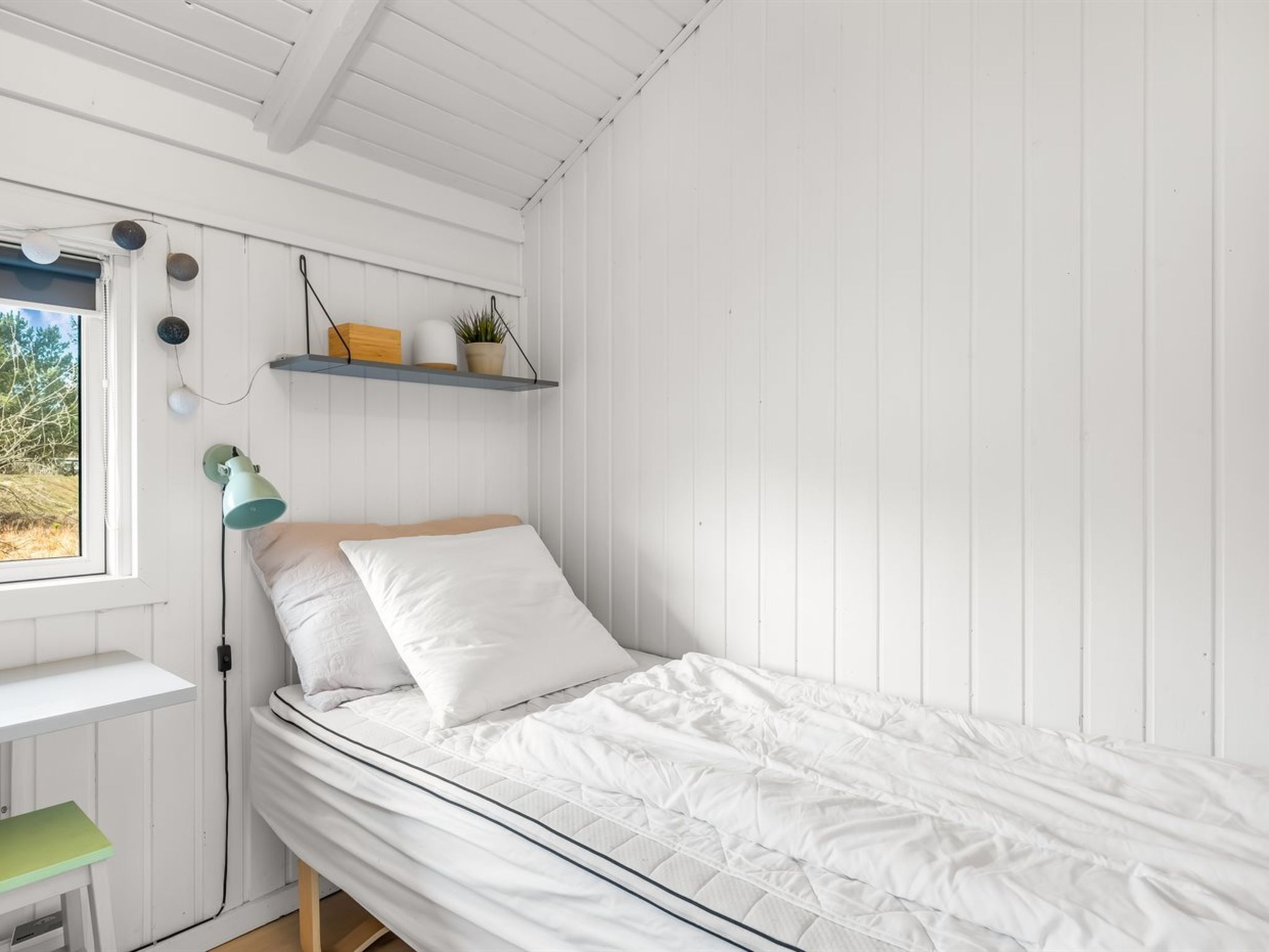 A single bed with white bedding, a green desk, a wall-mounted shelf with a plant, and a window with a view of trees.