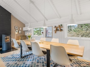 A modern dining area with a long table, white chairs, a wood-burning stove, and a large window with a view of trees. - "Julie" - 1.5km from the sea by Interhome (Fanø)