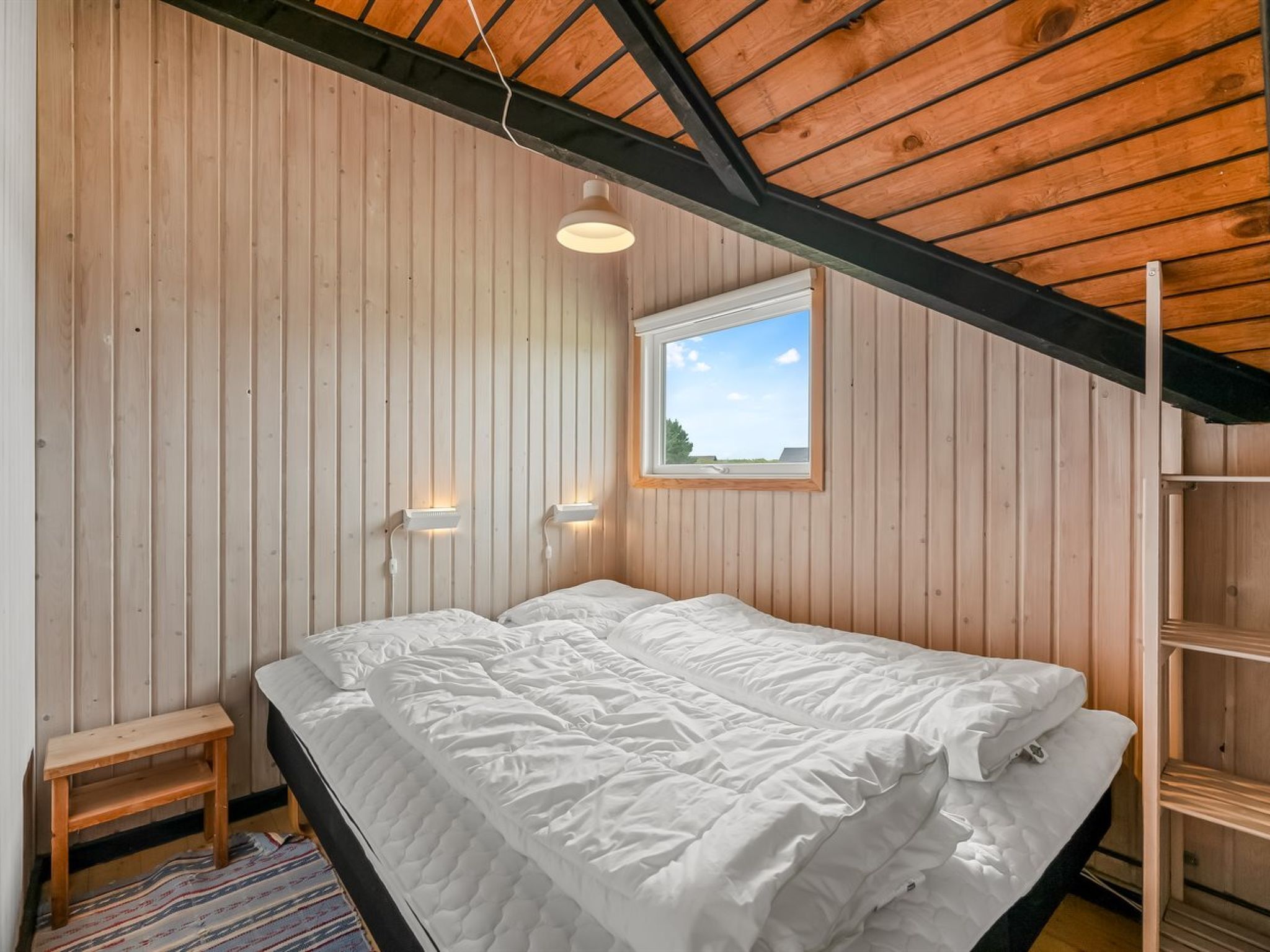 Une chambre avec un plafond en pente, des murs en bois, une fenêtre et un lit.