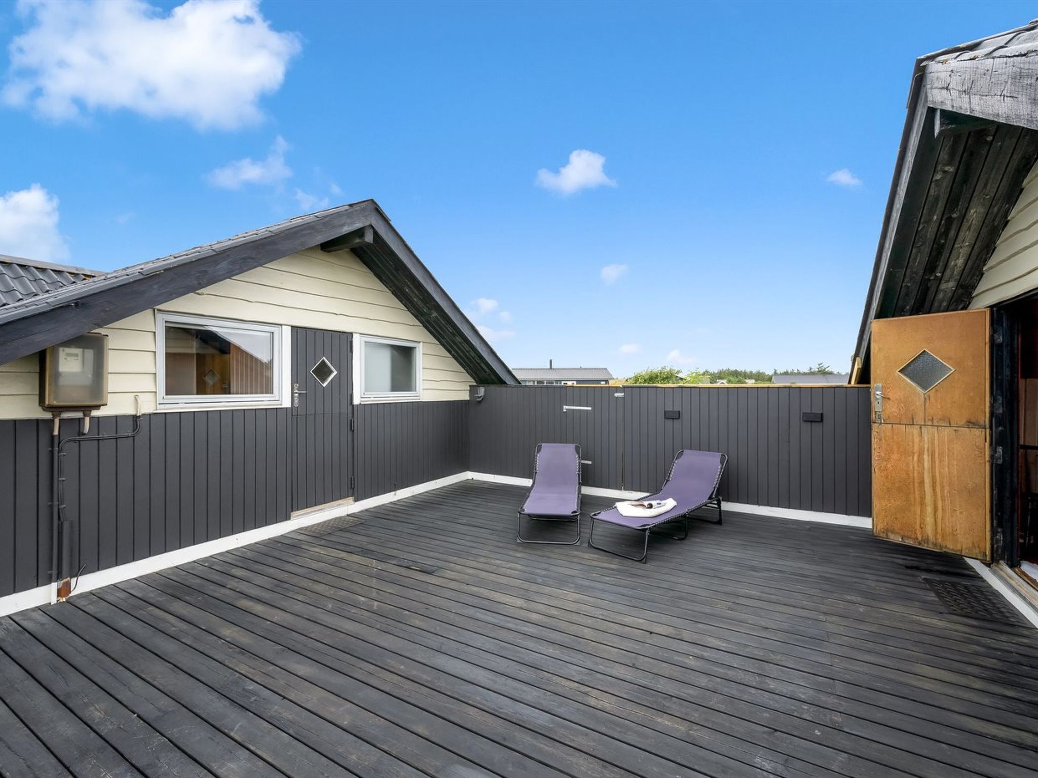Een houten terras met twee loungestoelen en een kleine tafel, omringd door een donkere schutting en een gebouw met een schuin dak.