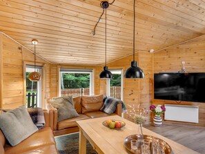 A cozy cabin interior with a wooden ceiling, leather sofa, a dining table with fruit and shot glasses, and a flat-screen TV.