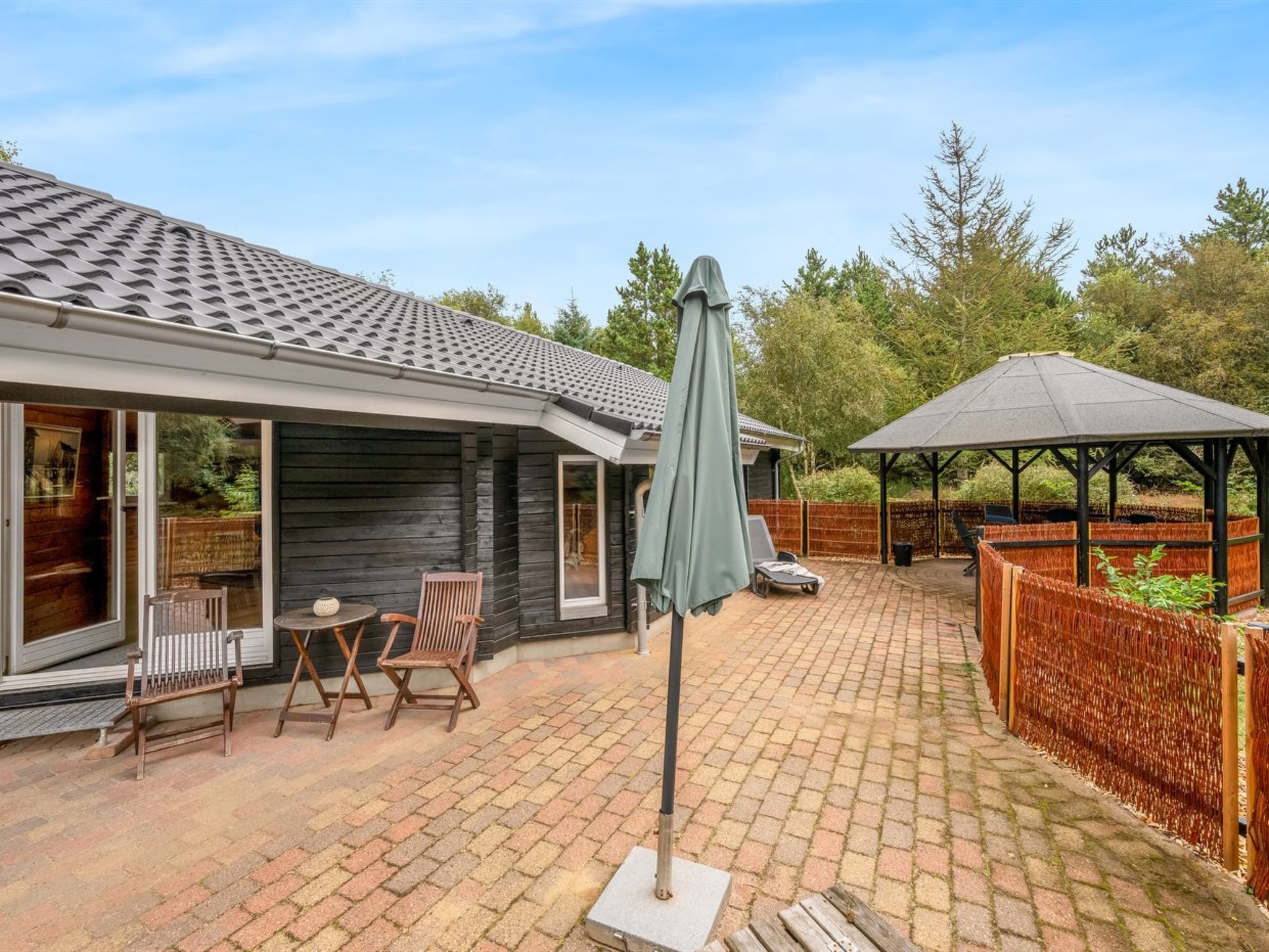 Un espace patio avec un sol en briques, deux chaises en bois, une table et un coin salon couvert avec une table.