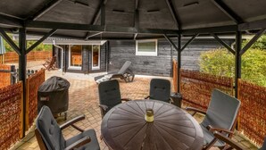 An outdoor patio with a round table and chairs, a grill, and a covered area with a view of a building. - "Jorina" - 700m to the fjord by Interhome (Ringkøbing)