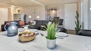 Een moderne woonkamer met een houten plafond, een eettafel gedekt met brood, een koffiepot en een potplant.