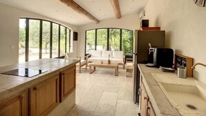 Cozinha moderna com uma ilha grande, armários de madeira e forno embutido. Há um espaço de estar com sofá e cadeiras, e janelas amplas com vista para o exterior.