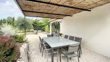 Een terras buiten met een tafel en stoelen, een pergola erboven en een tuin met planten en een pad.