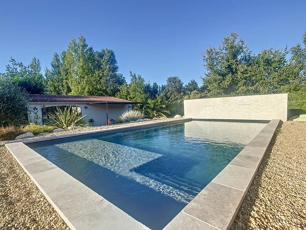 A rectangular swimming pool with clear water, surrounded by a pebble border, and a small building with a tiled roof in the background.