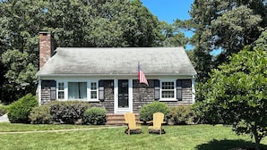 Une maison de plain-pied avec un porche à l’avant, une cheminée et deux fauteuils Adirondack sur la pelouse.