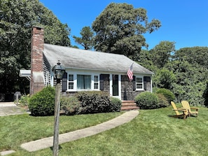 A single-story house with a brick chimney, a front porch with a bench, and a flagpole with an American flag.