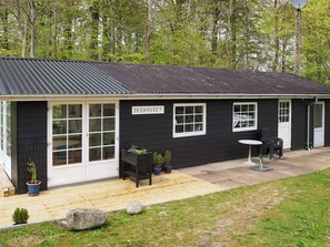 A small black building with a sign reading 'SKOVHUSET.' There are two windows, a door, and a small outdoor seating area with a table and chairs.