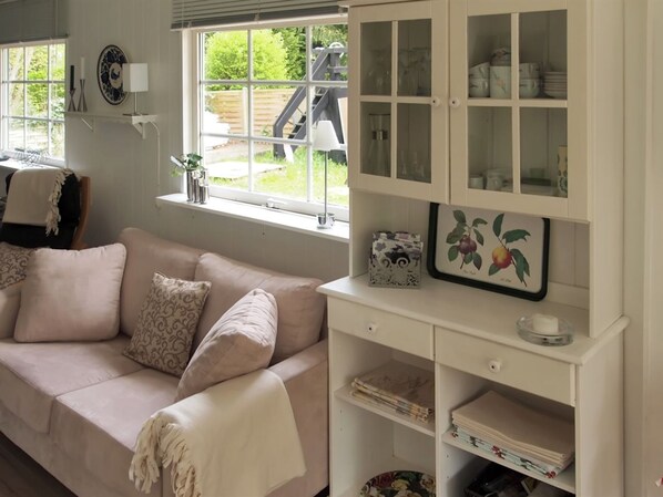 A cozy living room with a beige sofa, a white cabinet with glass doors, and a window with a view of a wooden deck and greenery.