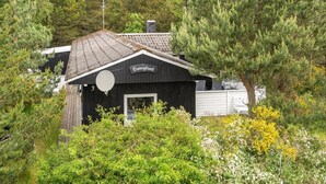 Een zwarte houten blokhut met een schotelantenne, omgeven door weelderige begroeiing en bomen.