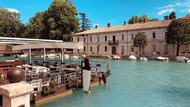 A riverside restaurant with outdoor seating, a stone building in the background, and boats docked along the water. - Fantastically beautiful ambience on 93sqm in Peschiera, 5 min=lake 7min=center, balcony (Peschiera del Garda)