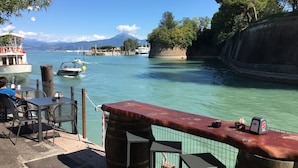 A waterfront restaurant with outdoor seating, a view of the water, and a mountain in the background. - Fantastically beautiful ambience on 93sqm in Peschiera, 5 min=lake 7min=center, balcony (Peschiera del Garda)