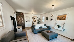 En moderne stue med en grå sofa, en blå sofa og et sofabord. I baggrunden er der et køkken med en hvid bordplade og en hængende plante.