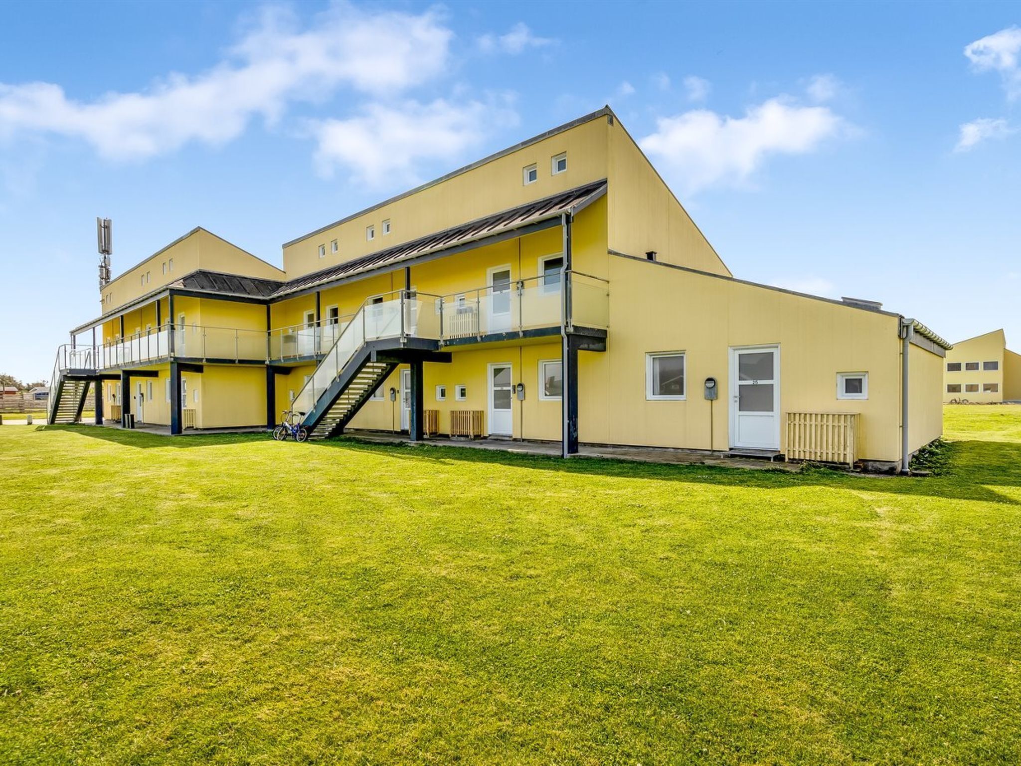 A yellow multi-story building with a staircase leading to a balcony, surrounded by a well-maintained lawn.