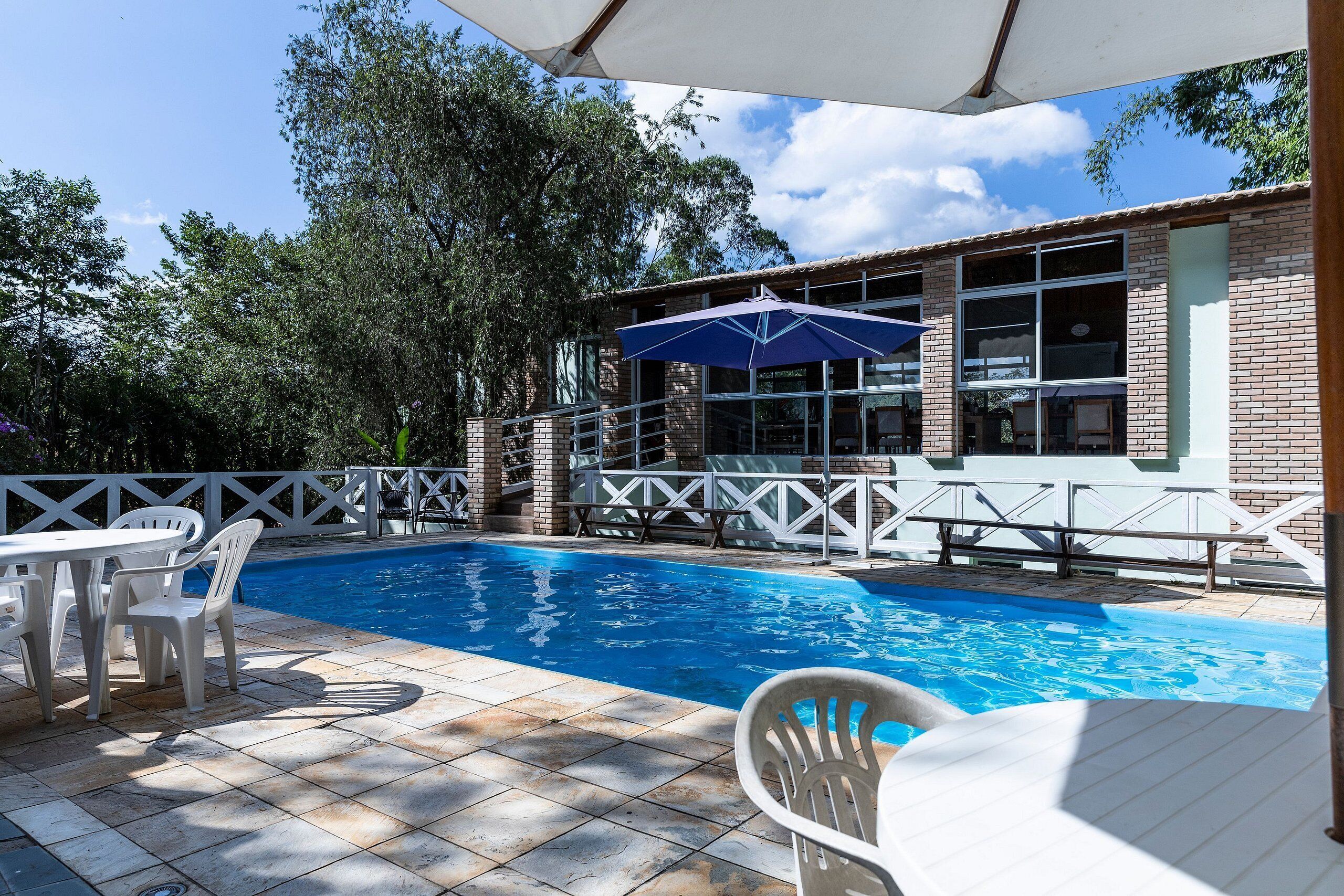 Piscina com deck azulejado, cadeiras brancas e guarda-sol azul.