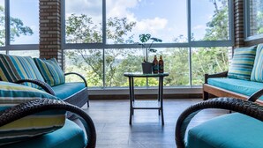 Une pièce meublée de mobilier en osier, avec une table en verre sur laquelle sont posées des bouteilles, et une vue sur les arbres à travers de grandes fenêtres.