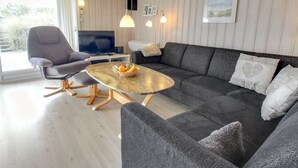 Un salon moderne avec un canapé gris, un fauteuil gris et une table basse ronde sur laquelle sont disposés des fruits. On y trouve également un téléviseur à écran plat, des lampes suspendues et une fenêtre avec des rideaux.