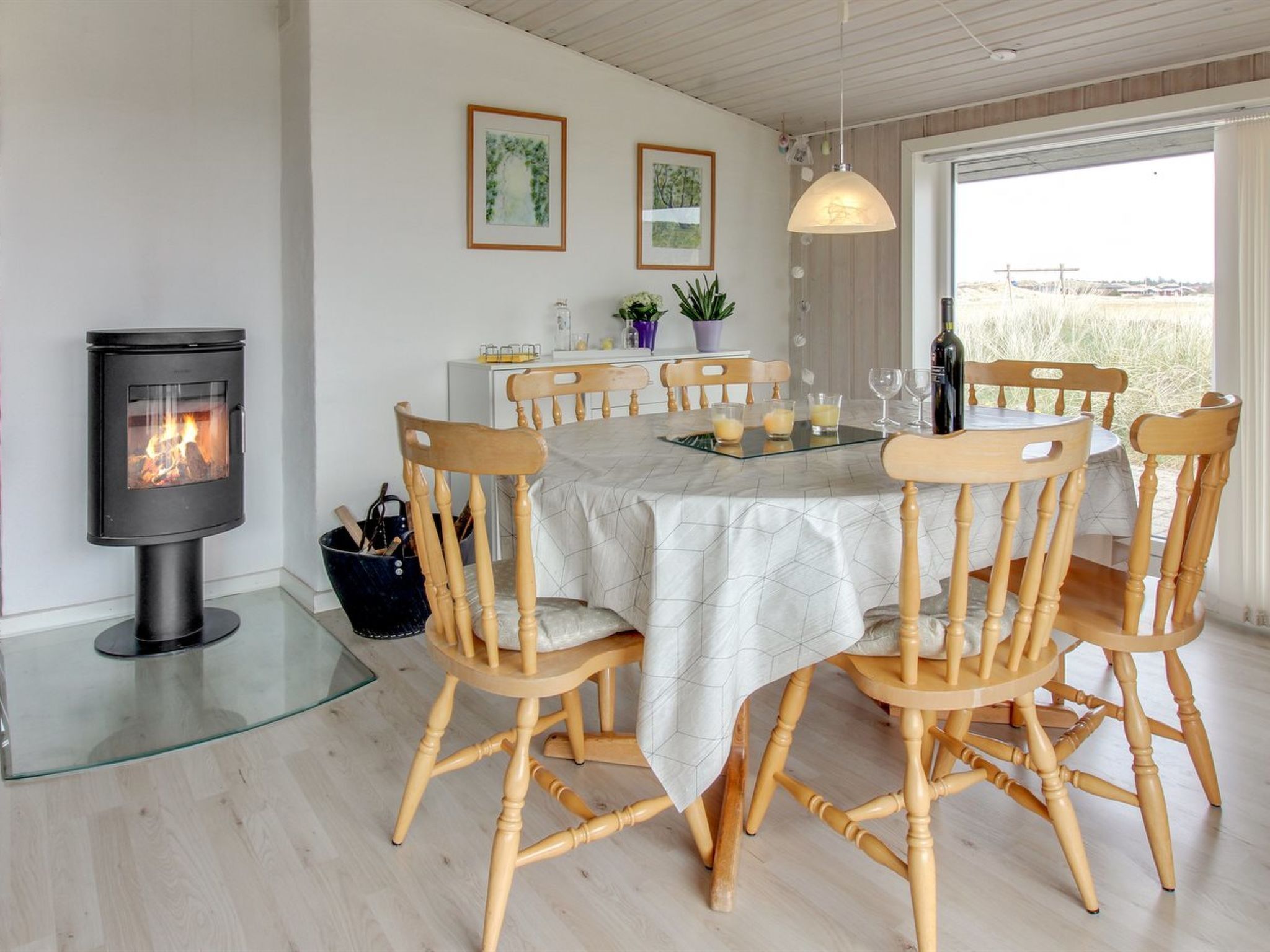 A dining area with a round table, wooden chairs, a glass-top table, a black wood-burning stove, and two framed pictures on the wall.