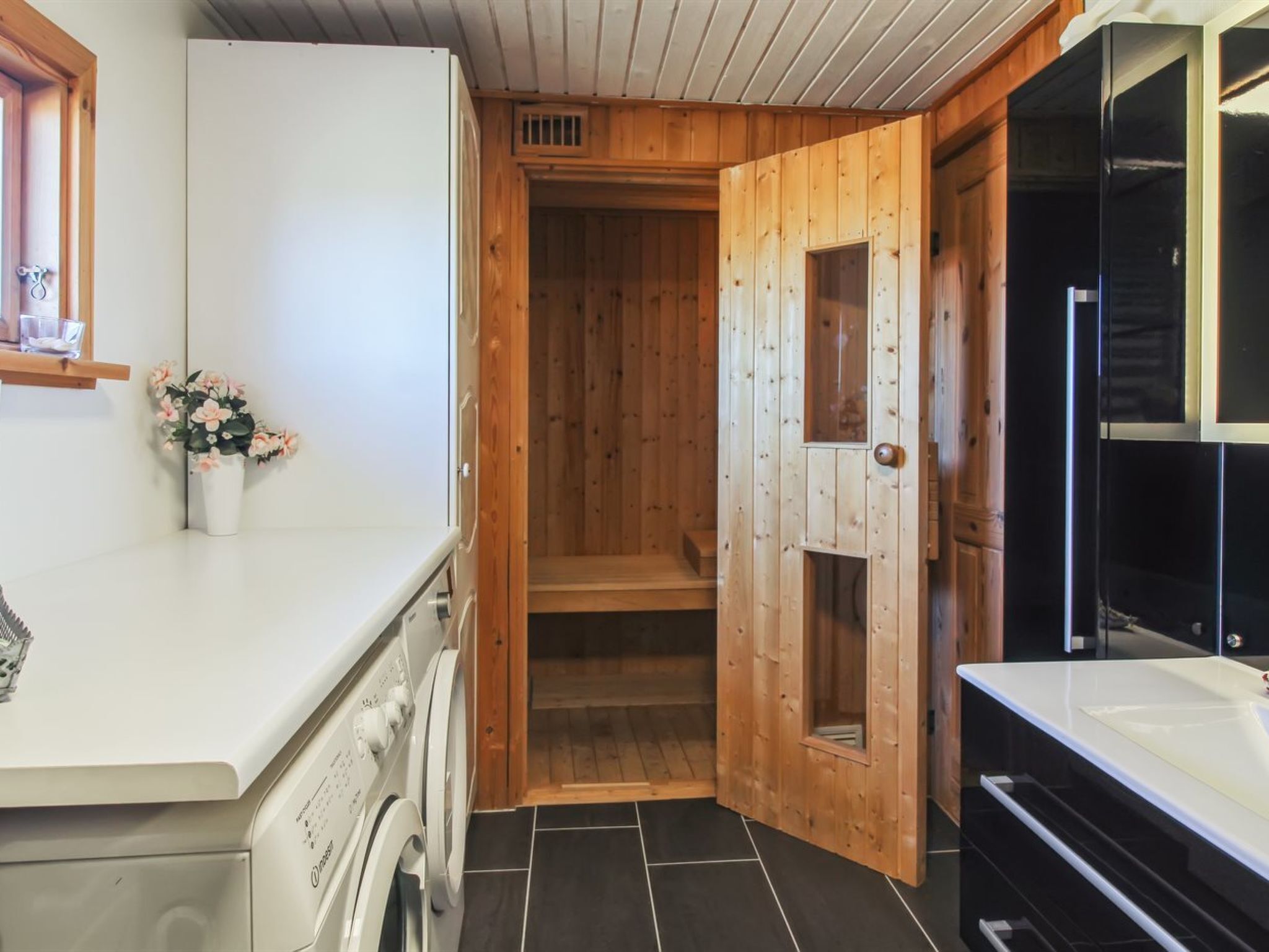 A compact kitchen with a built-in washing machine, wooden cabinets, and a small countertop with a vase of flowers.