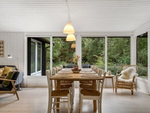 Un espace repas avec une table dressée pour six personnes, entourée de chaises, agrémenté d’un luminaire suspendu et offrant une vue sur la verdure grâce à de grandes fenêtres.