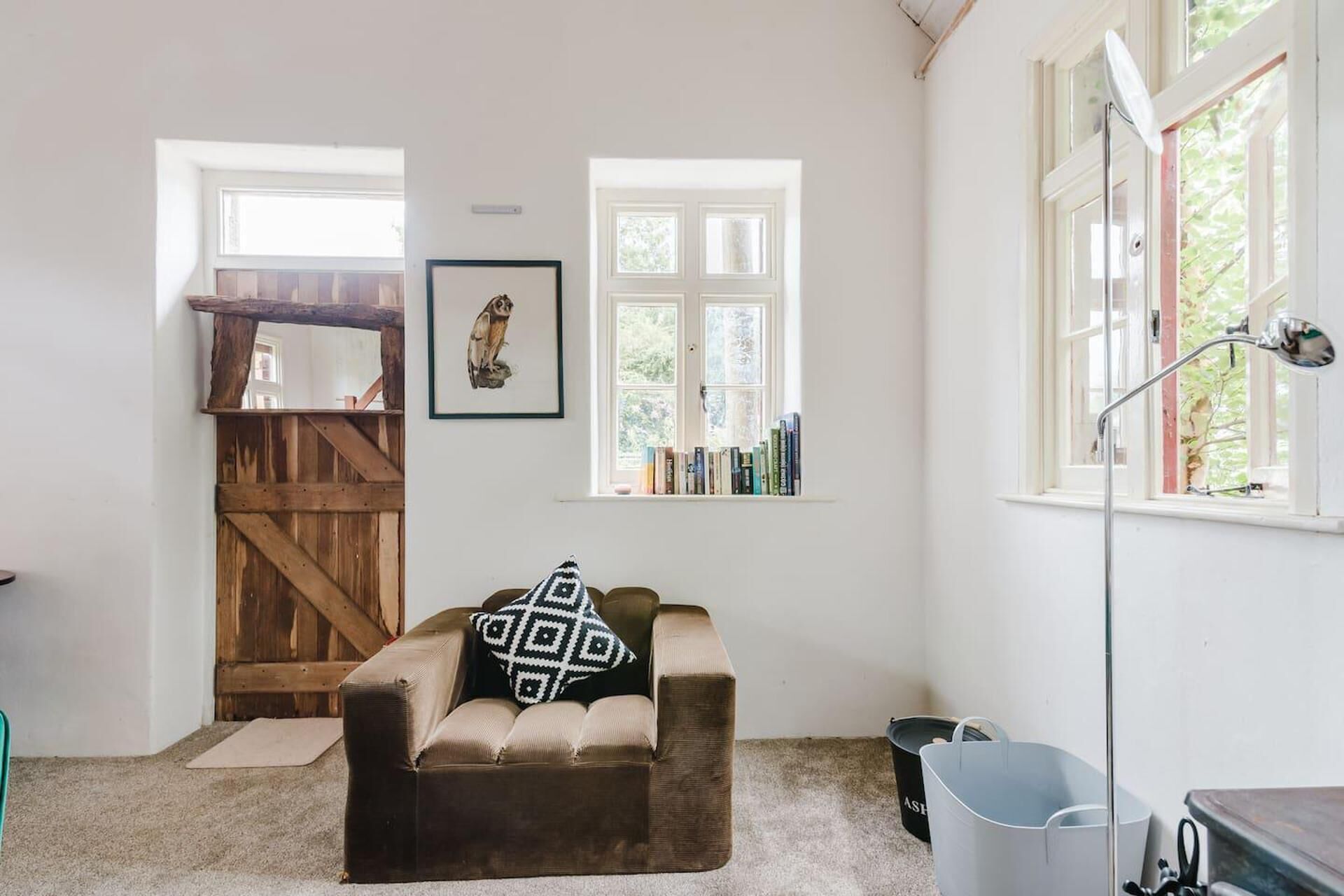 A cozy room with a brown sofa, a wooden door, a window with books, and a lamp.