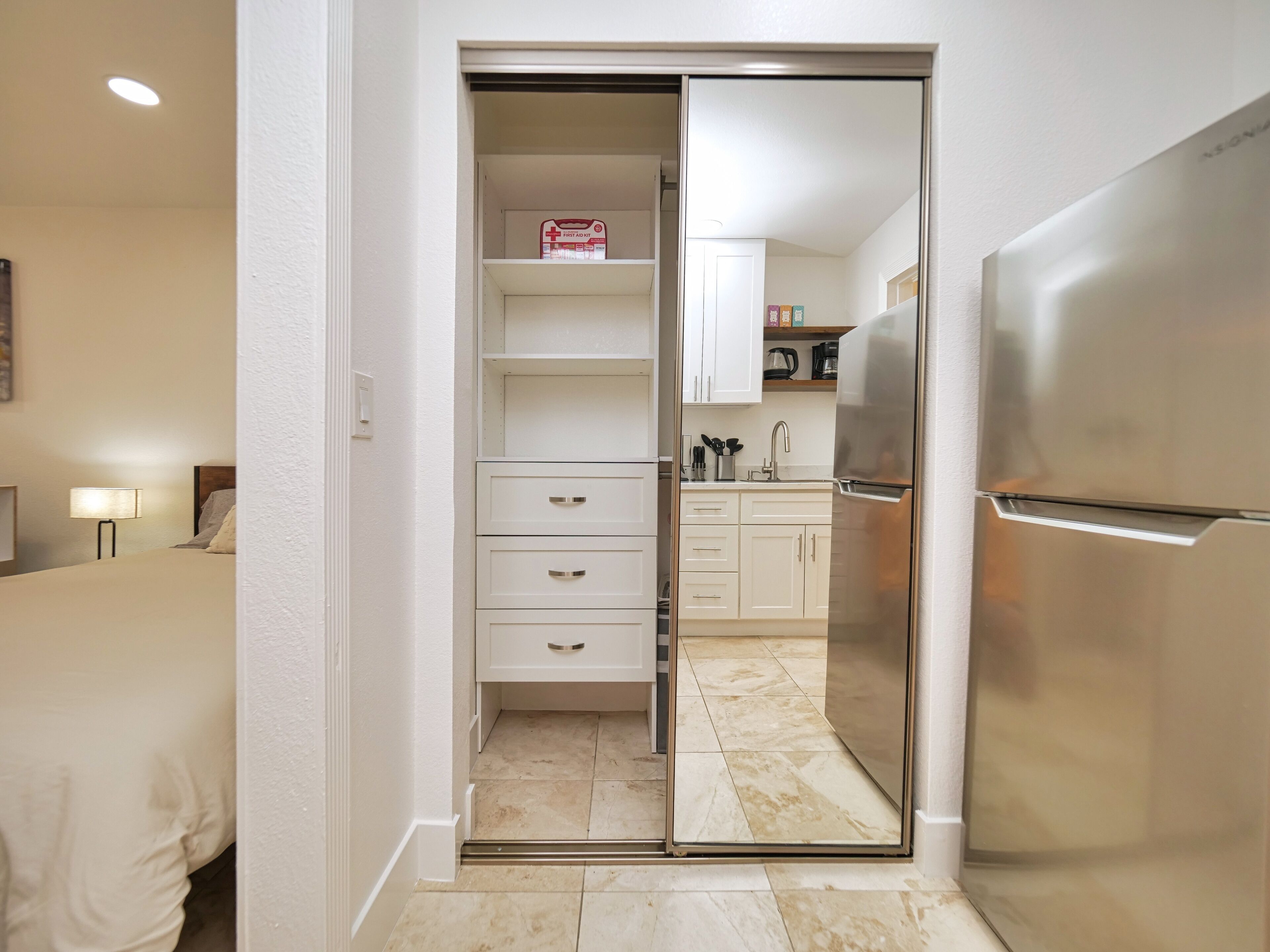 A hotel room with a bed, a nightstand, a lamp, a sliding door to a kitchenette, and a refrigerator.