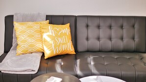 Una sala de estar moderna con un sofá de cuero oscuro, dos almohadones amarillos con texto blanco, una manta blanca, una mesa de centro de vidrio con un plato blanco y una alfombra estampada.