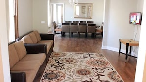 Un salon avec un canapé, une table à manger entourée de chaises, un tapis à motifs et un téléviseur fixé au mur.