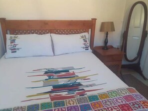 Ein Bett mit weißer Bettdecke und bunten geometrischen Mustern, ein hölzernes Kopfteil, ein Nachttisch mit Lampe und ein Spiegel.