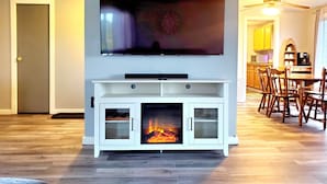 Una sala de estar con un televisor de pantalla plana montado sobre un mueble blanco, una chimenea, piso de madera y un comedor con mesa y sillas.