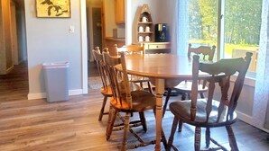 Un comedor con una mesa y sillas de madera, una cafetera y vistas a la vegetación a través de amplias ventanas.