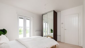 A bedroom with a bed, a large mirror, a wardrobe, and a window. - 5 Bed House - Sleeps 9 - Pets - Garden - Parking (Ashford)