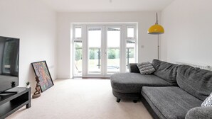 A modern living room with a grey sofa, a black TV stand, a framed artwork, and a yellow pendant light. - 5 Bed House - Sleeps 9 - Pets - Garden - Parking (Ashford)
