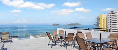 A rooftop terrace with a glass balustrade, a table with chairs, and a view of the ocean and distant islands.