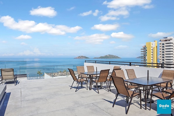 A rooftop terrace with a glass balustrade, a table with chairs, and a view of the ocean and distant islands.