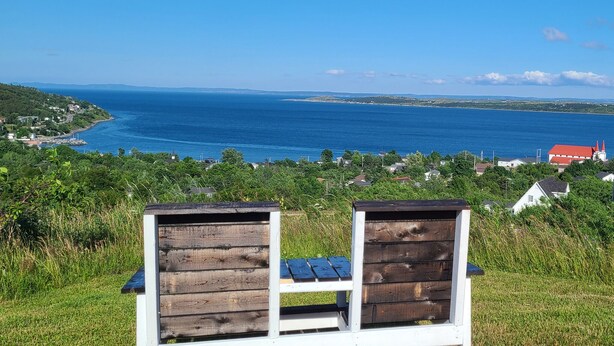 A wooden bench with a view of a coastal landscape, including a body of water and distant buildings. - Spaniard's Perch - Incredible Ocean View (Spaniard's Bay)