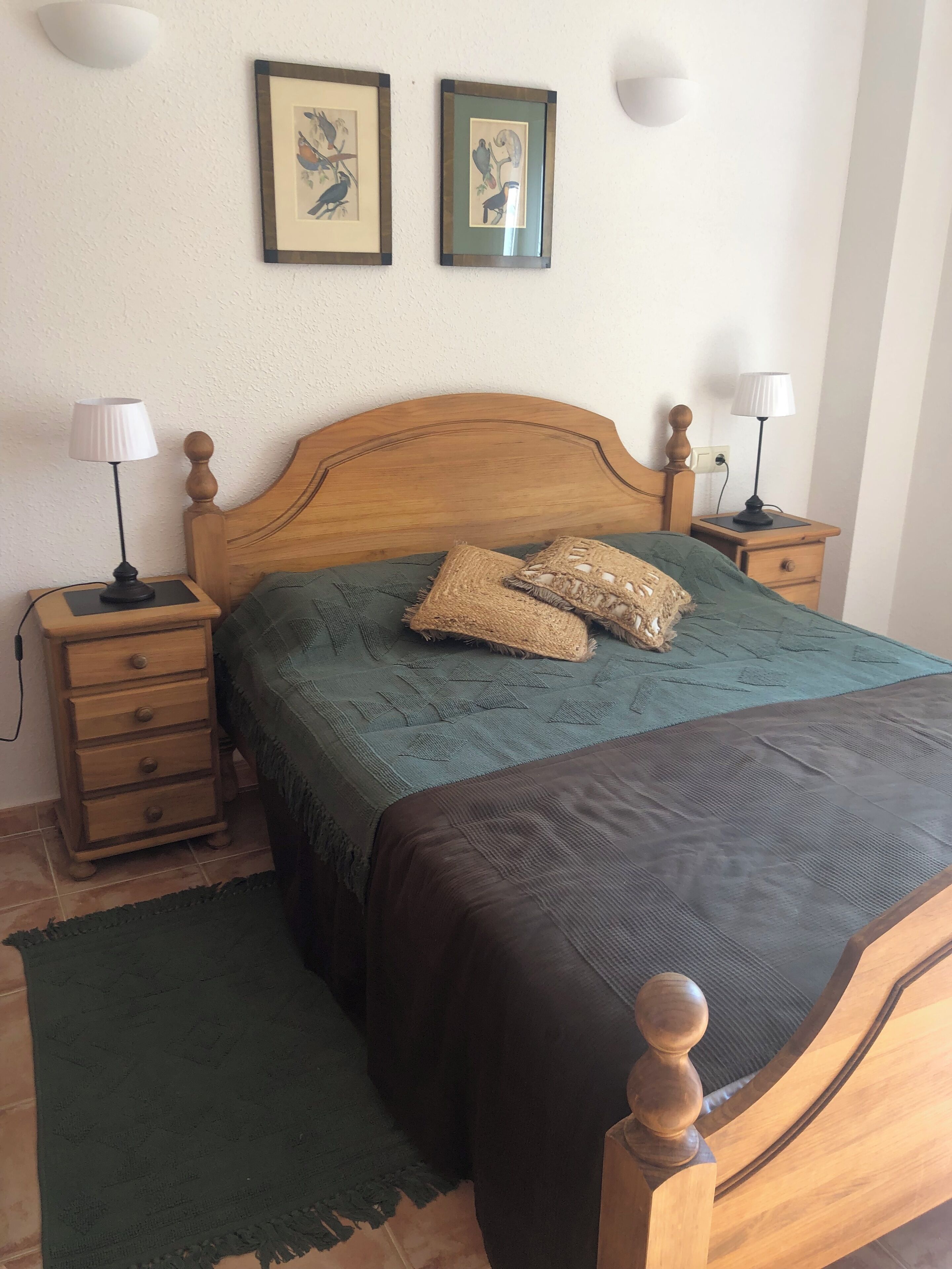 Una cama bien hecha con cabecera de madera, dos mesitas de noche con lámparas, una manta verde y dos cuadros en la pared.
