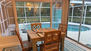 A wooden dining table with chairs, a checkered placemat, and a view of a swimming pool through large glass doors. - Coco NoKo cozy beach cottage + Swimming pool! Golf cart and pet friendly! (Nokomis)
