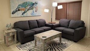 A living room with a sectional sofa, a coffee table, a floor lamp, a painting of a sea turtle, and a small side table with a decorative item. - Coco NoKo cozy beach cottage + Swimming pool! Golf cart and pet friendly! (Nokomis)
