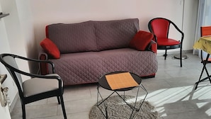 Un espace de vie comprenant un canapé gris, deux fauteuils rouges, une petite table ronde et une nappe à motifs.