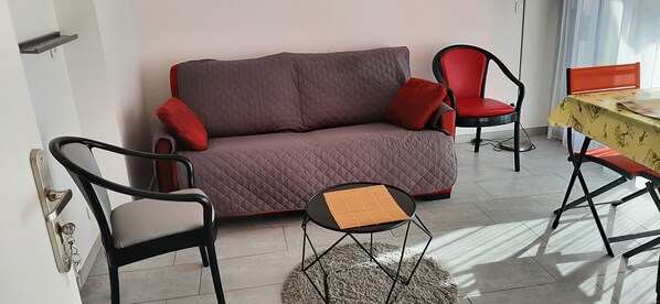 A living area with a grey sofa, two red chairs, a small round table, and a patterned tablecloth.
